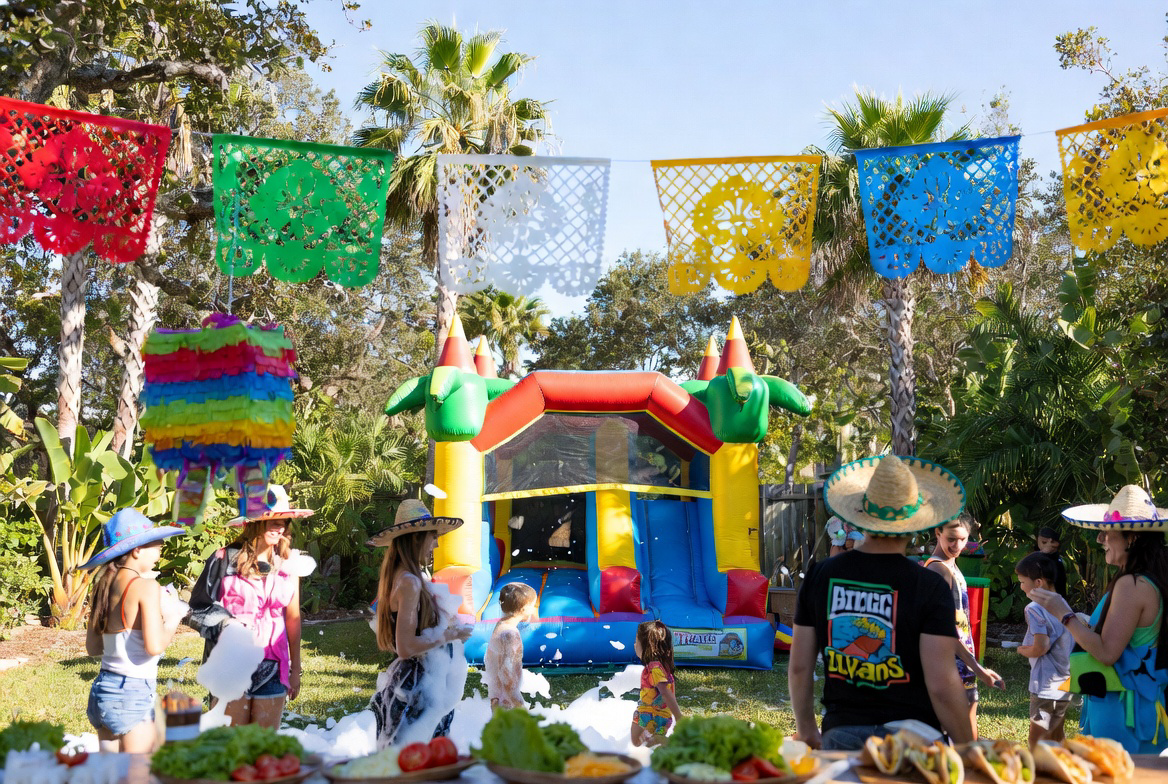 Festive backyard Cinco de Mayo party with colorful bounce house, papel picado banners, taco bar, and string lights at sunset